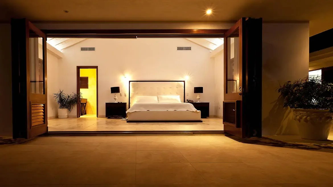 A beautifully styled bedroom opens through folding wooden doors, blending indoor comfort with the villa's outdoor tropical surroundings. Warm ambient lighting adds a calm, intimate tone suited to a high-end Anguilla stay.