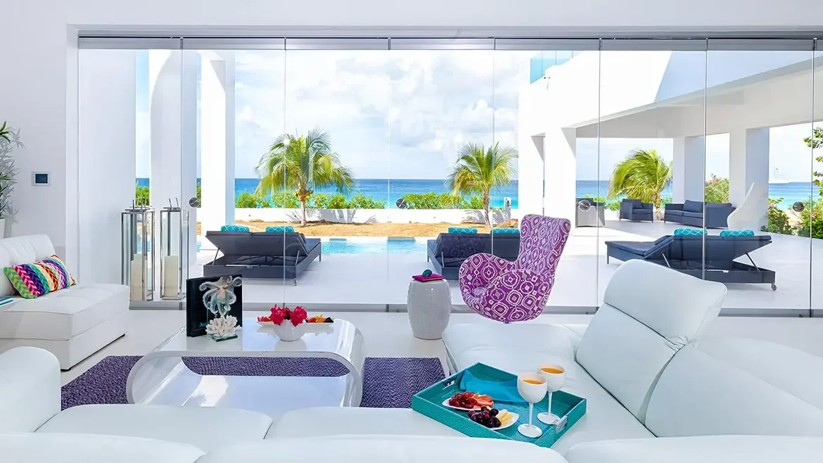 Experience unparalleled luxury at this stunning Anguilla beach house, featuring expansive floor-to-ceiling glass walls that showcase breathtaking Caribbean Sea views. The modern interior blends sleek white furnishings with vibrant pops of color, offering a seamless transition to the private pool and pristine white sands.