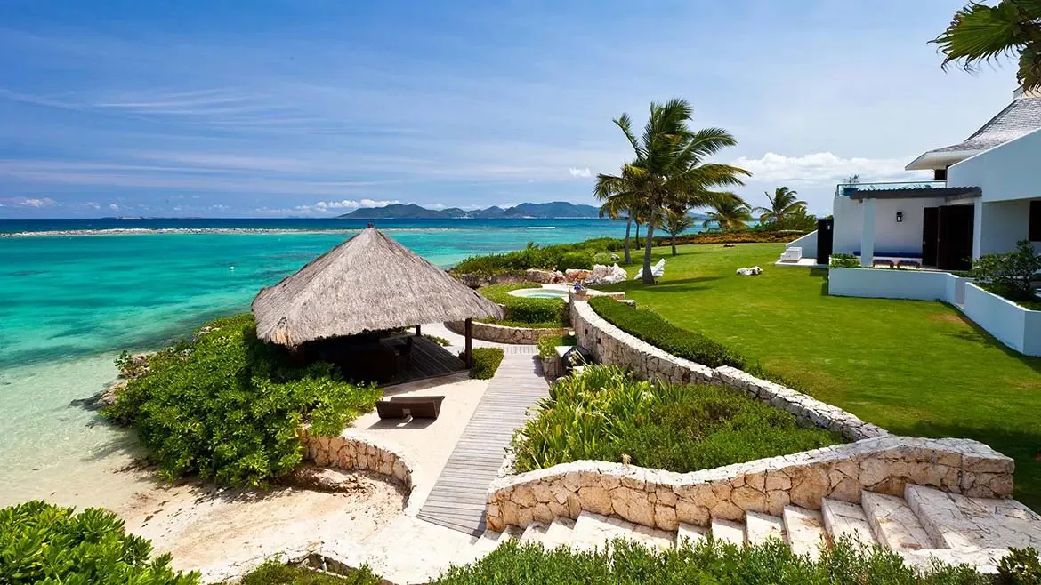 The stone terrace and thatched gazebo at Le Bleu Villa overlook the sea, offering a private vantage point for lounging or entertaining. Bright daylight and lush landscaping enhance the villa's resort-style outdoor appeal.