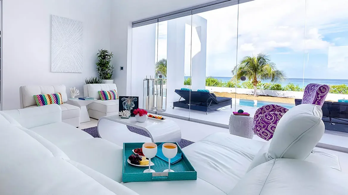 This ultra-modern luxury villa in Anguilla features an expansive open-concept living area with floor-to-ceiling glass walls that showcase breathtaking Caribbean Sea views. The pristine white interior is accented with vibrant tropical decor, leading out to a private infinity pool and sun-drenched terrace for the ultimate island retreat.