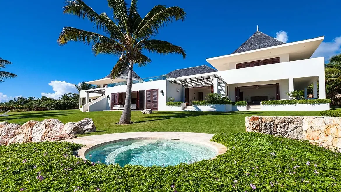A circular outdoor spa anchors this sunlit lawn view, with modern villa architecture and a mature palm tree completing the composition. The image emphasizes Le Bleu Villa's expansive grounds and private resort amenities.