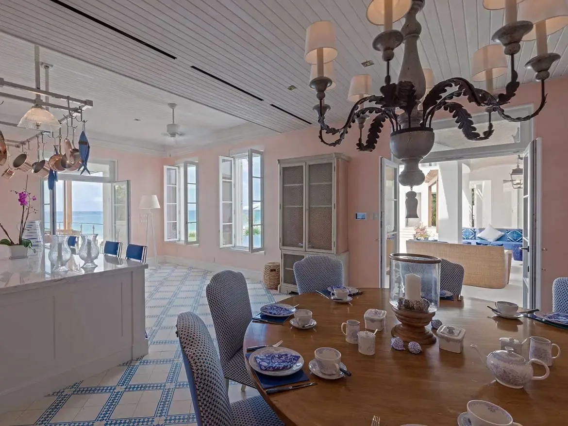 This elegant dining space at Cerulean Villa features soft pink walls, a large wooden table set for tea, and panoramic views of the Caribbean Sea. The airy interior design blends coastal charm with luxury, offering seamless indoor-outdoor living through expansive white-shuttered windows.