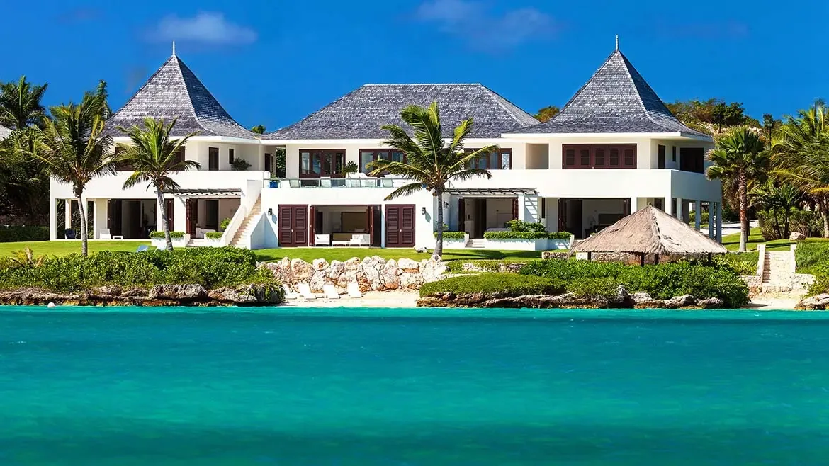 This exterior perspective showcases Le Bleu Villa's white Caribbean architecture set above vivid blue water and lush tropical landscaping. The bright, clean composition is ideal for presenting the property's oceanfront prestige in Anguilla.