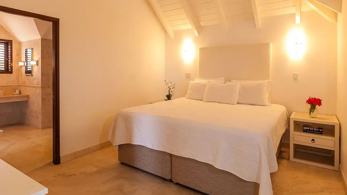 This serene bedroom suite at Villa Indigo features a plush king-sized bed, soft ambient lighting, and elegant minimalist decor. The spacious layout includes a private ensuite bathroom and high vaulted ceilings, offering a tranquil retreat for a luxury Anguilla vacation.