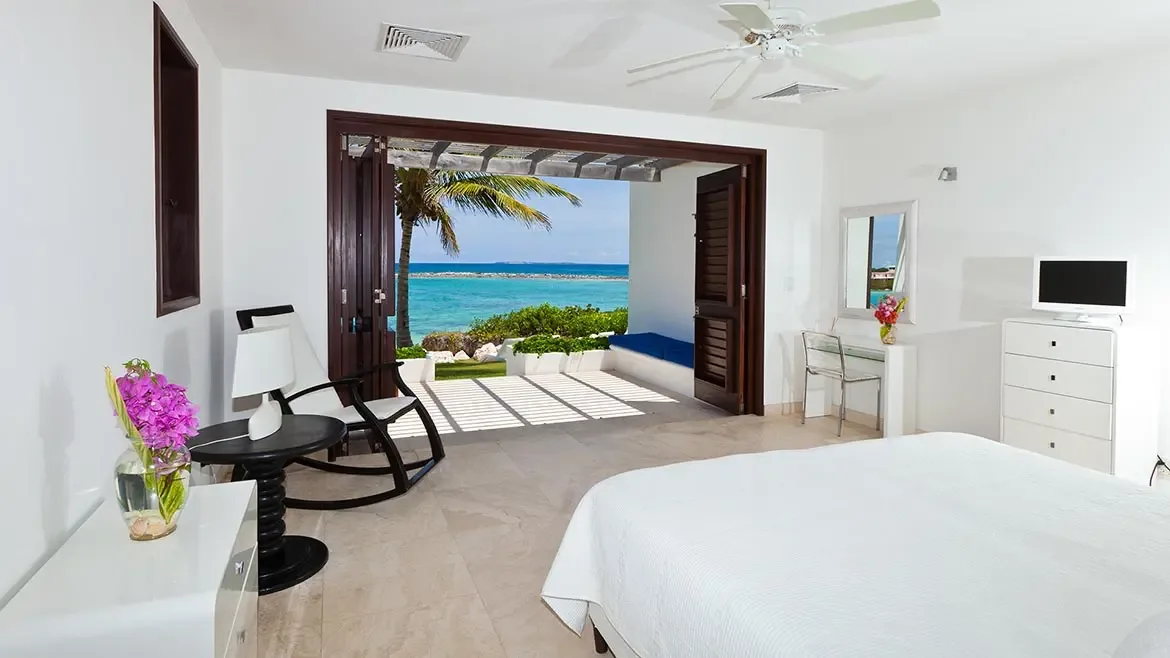 This serene bedroom opens directly to a private oceanfront patio, where turquoise waters and a swaying palm complete the view. Natural light and understated furnishings give the suite an elegant, restful island feel.