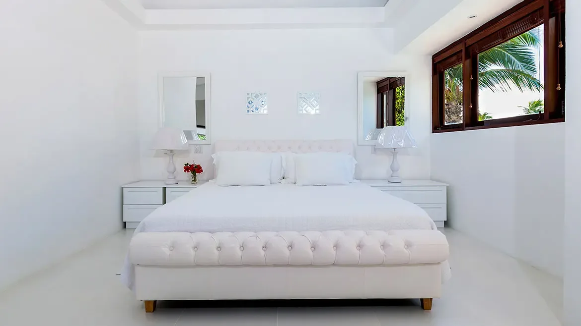Experience pure tranquility in this elegant all-white suite at Villa Indigo, a premier luxury rental in Anguilla. The sophisticated bedroom features a plush tufted bed and minimalist decor, offering a serene sanctuary with beautiful tropical views through expansive windows.