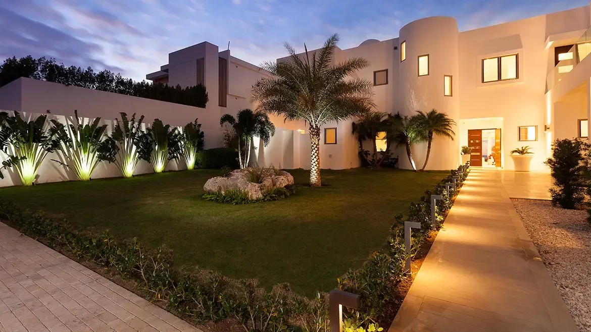 This ultra-modern luxury villa in Anguilla features a stunning illuminated landscape with manicured lawns and tropical palm trees. The white minimalist architecture creates a striking contrast against the evening sky, offering an elegant sanctuary for a high-end Caribbean escape.
