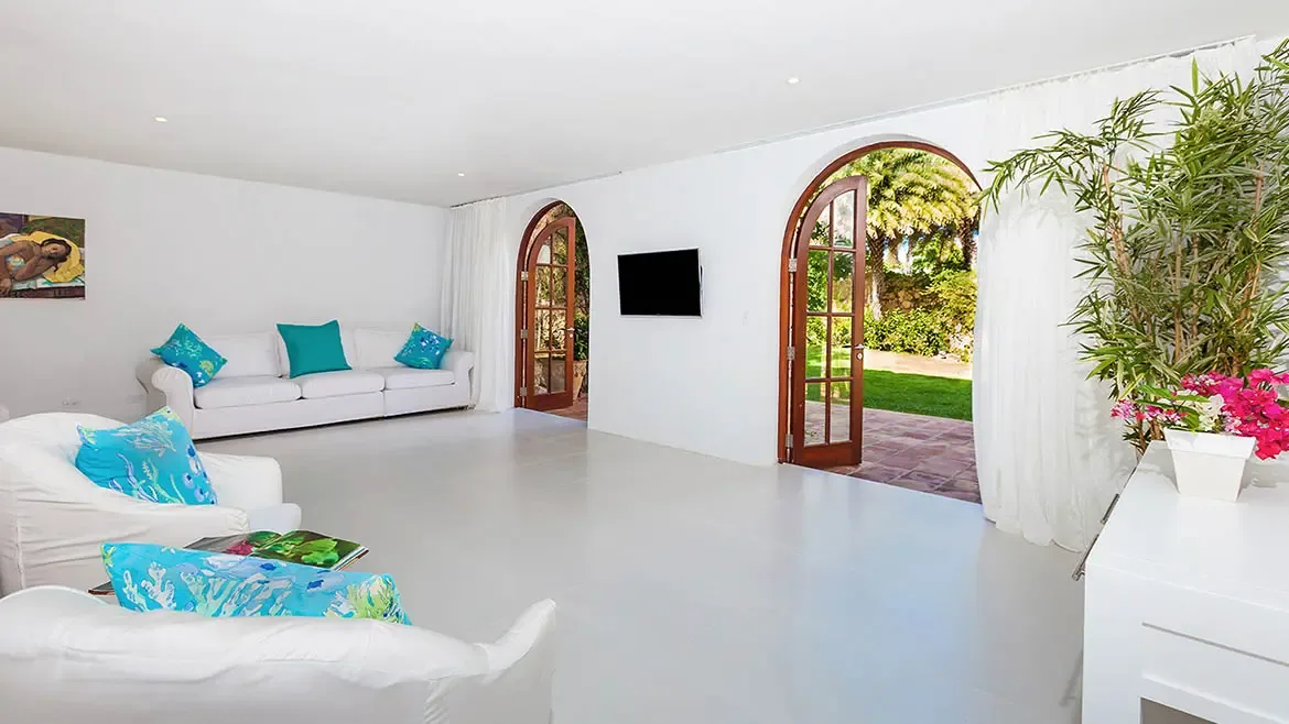 This expansive, bright living area at Villa Indigo in Anguilla features sleek white decor accented by vibrant turquoise pillows and tropical greenery. The arched French doors open directly onto a lush garden terrace, offering a seamless indoor-outdoor lifestyle for a luxury Caribbean escape.