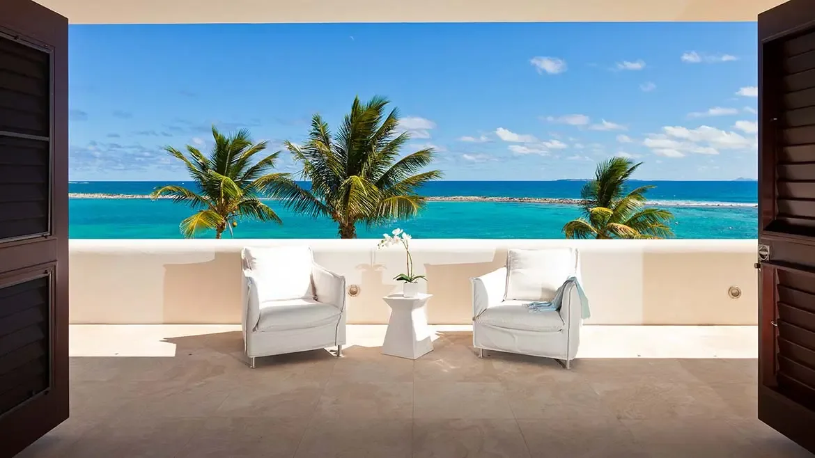 Elegant white armchairs on a stone-tiled balcony create a quiet vantage point over the turquoise coastline. This image adds a refined lifestyle moment to the Le Bleu Villa gallery while showcasing the property's scenic outdoor spaces.