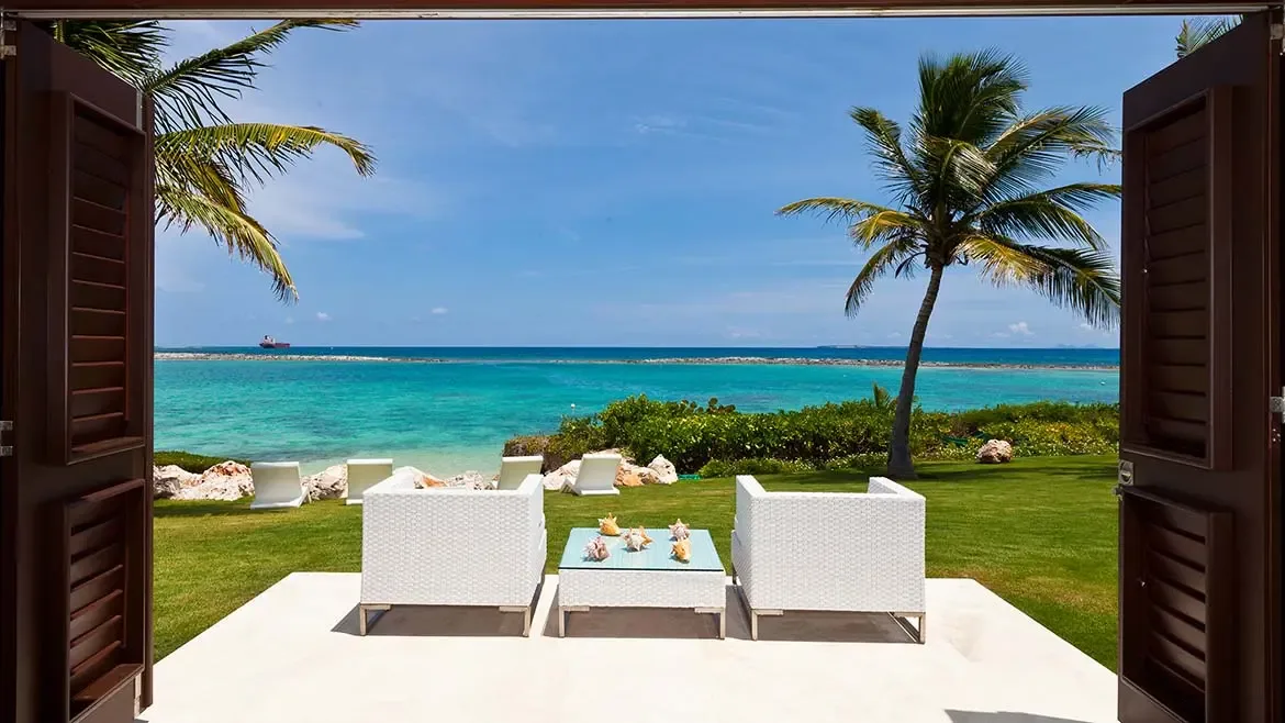 Open louvered doors reveal a furnished patio, green lawn, and brilliant ocean beyond, emphasizing the villa's seamless indoor-outdoor flow. This perspective is especially effective for conveying the relaxed elegance of Le Bleu Villa.