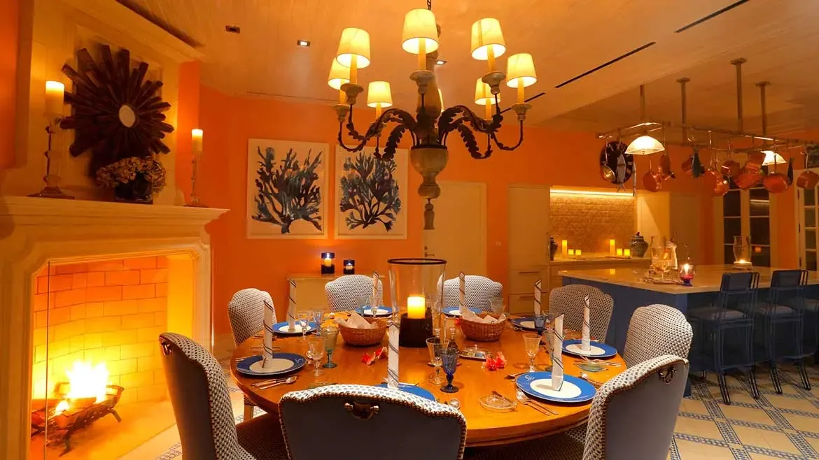 This sophisticated dining space at Cerulean Villa features a warm, inviting ambiance highlighted by a glowing fireplace and an ornate chandelier. Perfect for hosting elegant dinner parties, the room blends coastal blue accents with vibrant orange walls to create an unforgettable Caribbean atmosphere.
