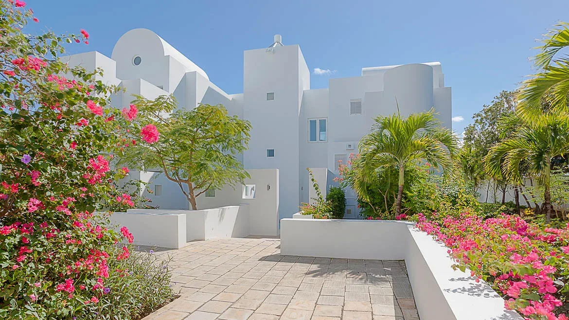 Paved courtyard and manicured gardens with bright pink flowers surrounding the white architecture of the villa.