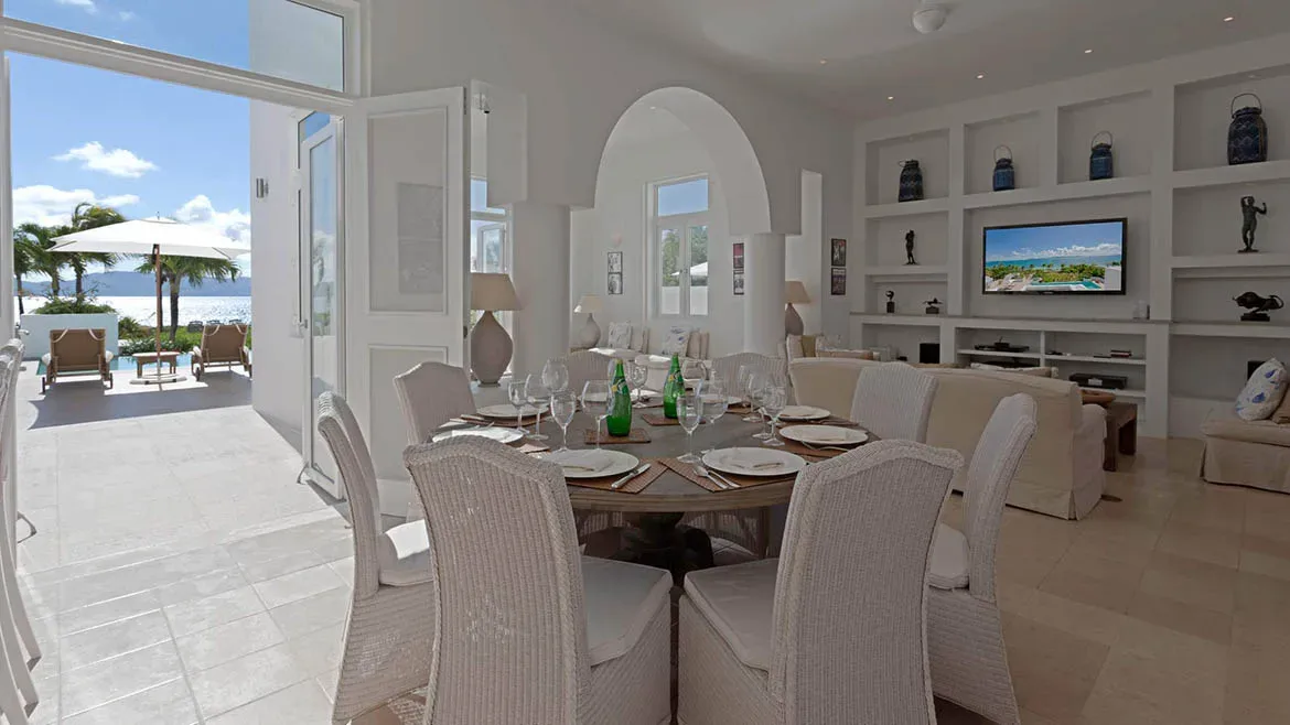 An airy white dining room with a round table set for guests, opening directly onto a sunny terrace with ocean views.