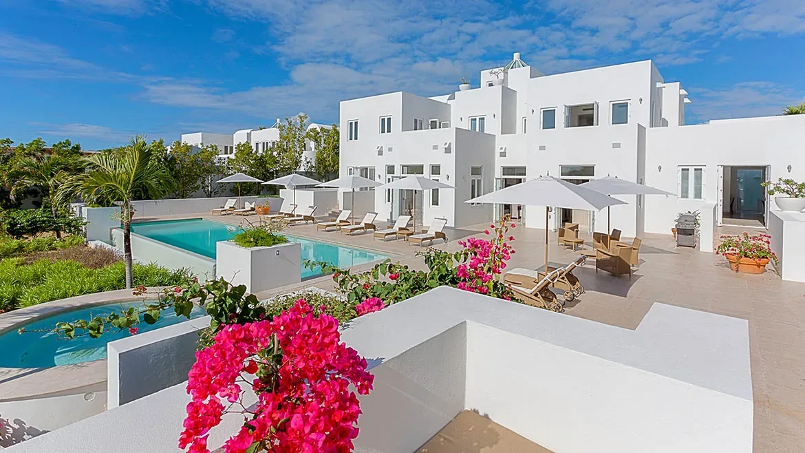 View of the white villa and swimming pool framed by vibrant pink bougainvillea flowers.