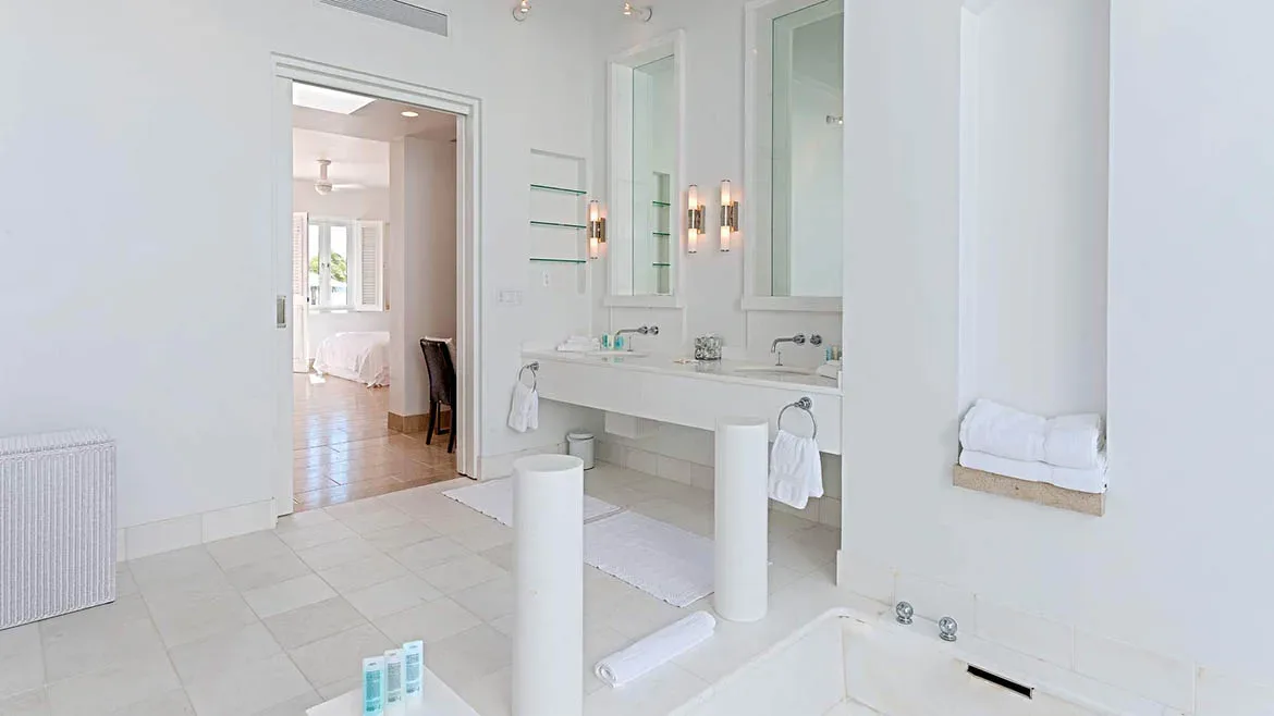 Bright, spacious white luxury bathroom with marble counters and a view into the bedroom.
