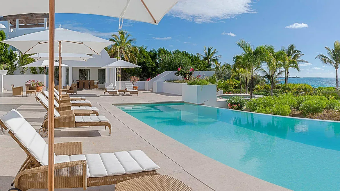 Luxury swimming pool with white sun loungers and umbrellas overlooking the turquoise ocean and palm trees.