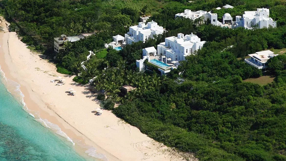 Aerial view of white luxury villas surrounded by lush green tropical foliage next to a white sand beach and turquoise ocean.