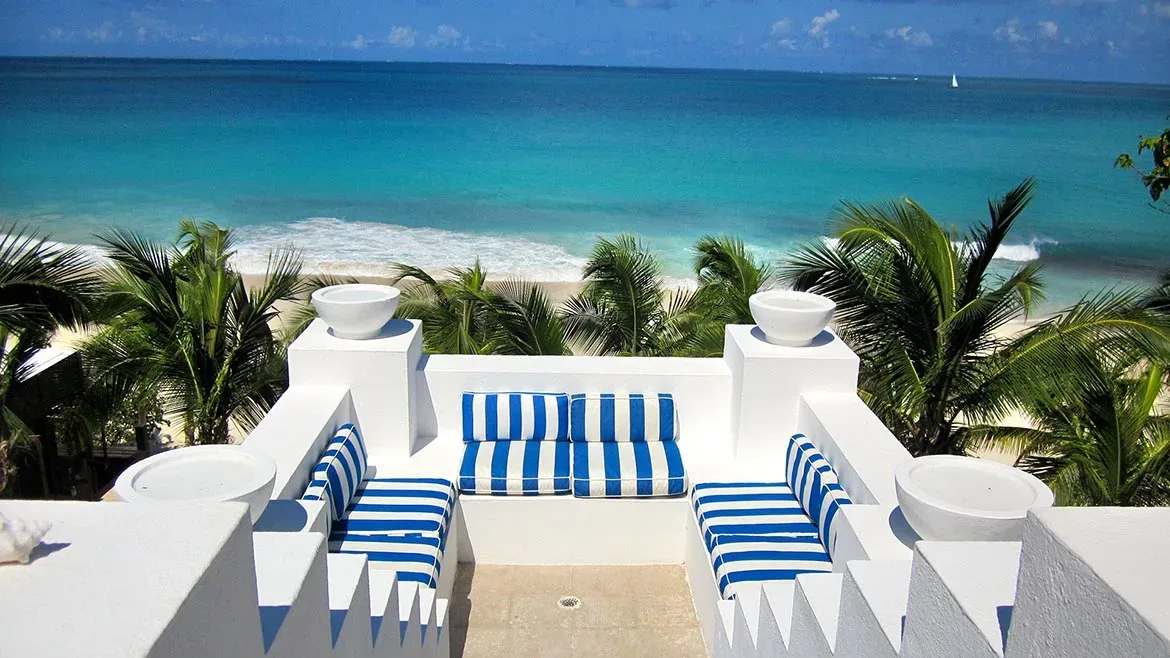A white built-in outdoor seating area with blue and white striped cushions overlooking the turquoise ocean and palm trees.