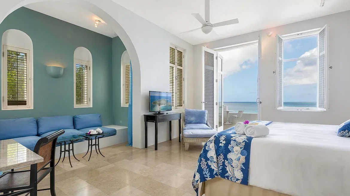 Bright bedroom with white walls, blue accents, and wooden shutters overlooking the sea.