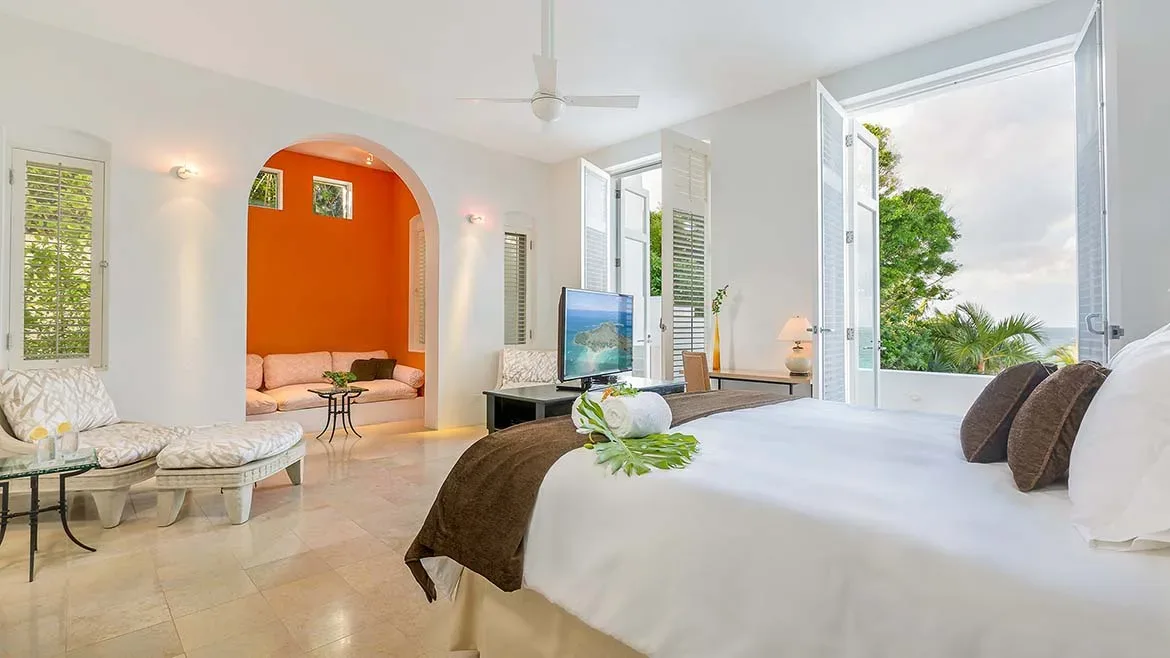 Bright bedroom interior featuring a white bed, open shutters leading to the ocean, and a cozy orange alcove.