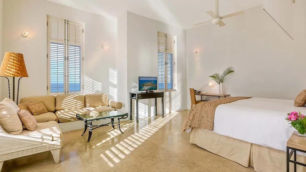 Sunny bedroom suite with neutral tones, comfortable seating, and sunlight filtering through window shutters.