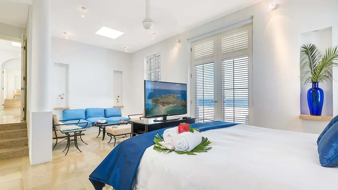 Modern bedroom with white decor, a blue bed runner, and a flat-screen TV.