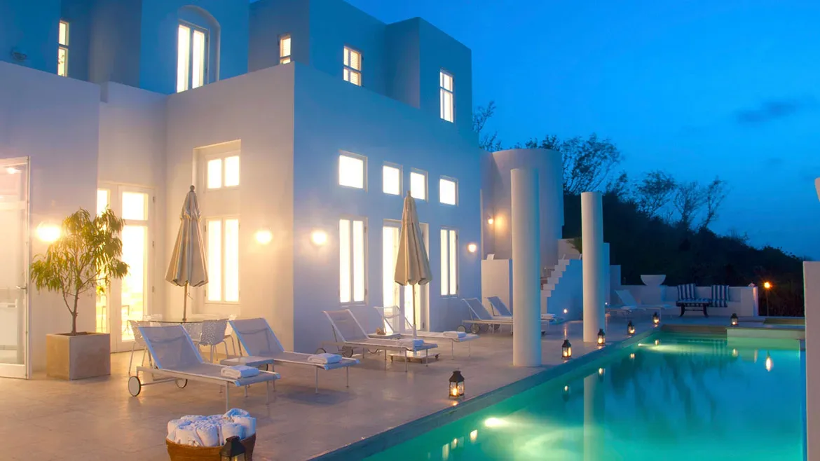 Evening view of the illuminated white villa and swimming pool with loungers under a deep blue twilight sky.