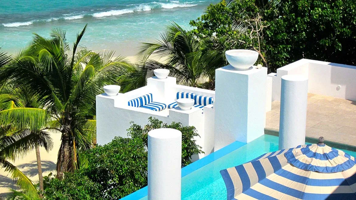 High-angle view of an infinity pool with a striped umbrella and white architectural pillars overlooking the beach.