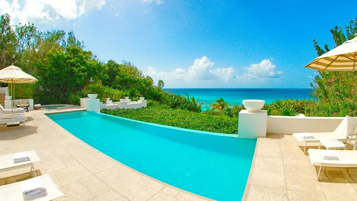 A turquoise infinity pool with white loungers and towels, overlooking lush green gardens and the blue Caribbean Sea.