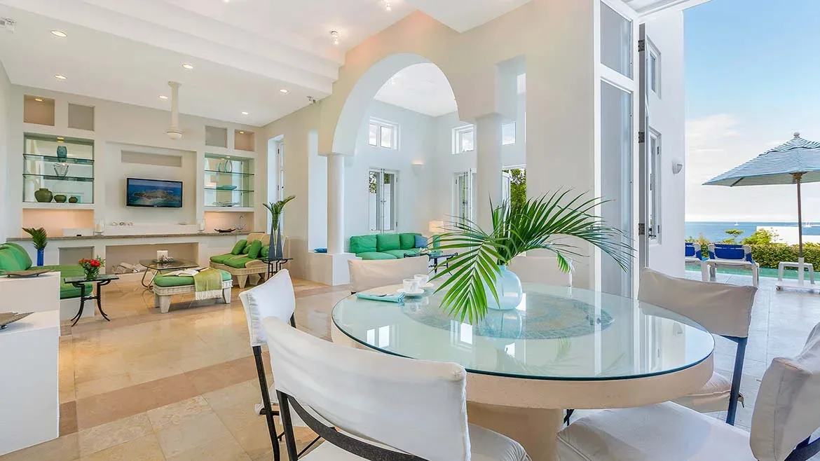 White interior with a glass dining table, green sofas, and an archway leading to a bright living area.