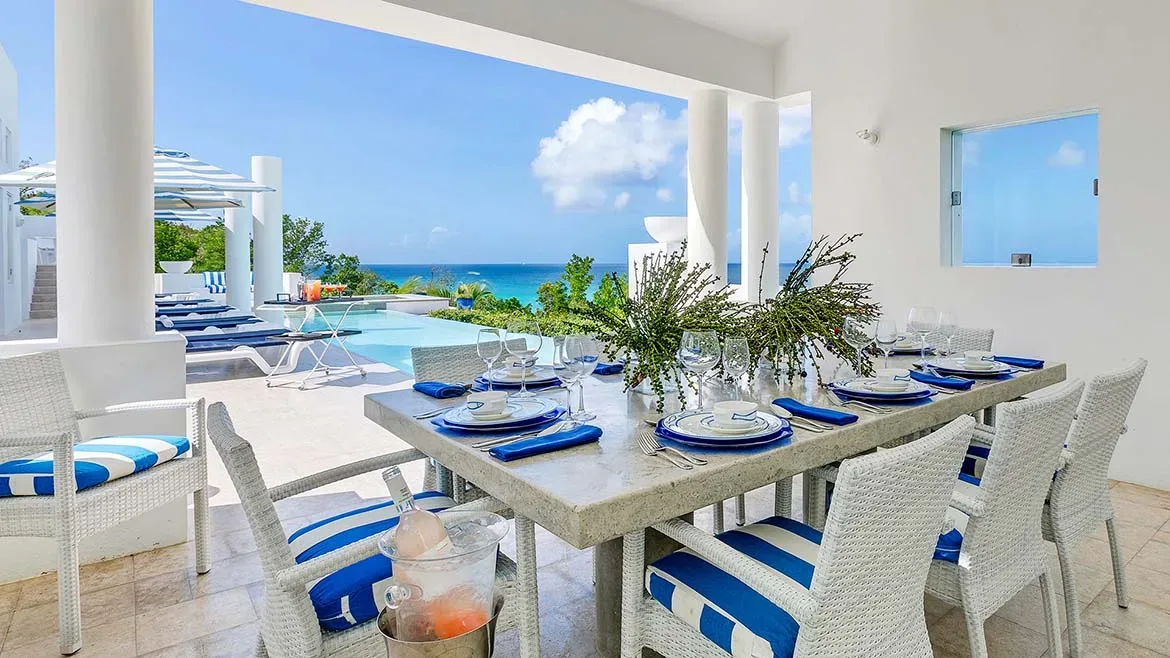 Outdoor dining table set for a meal on a white patio overlooking a swimming pool and the ocean.