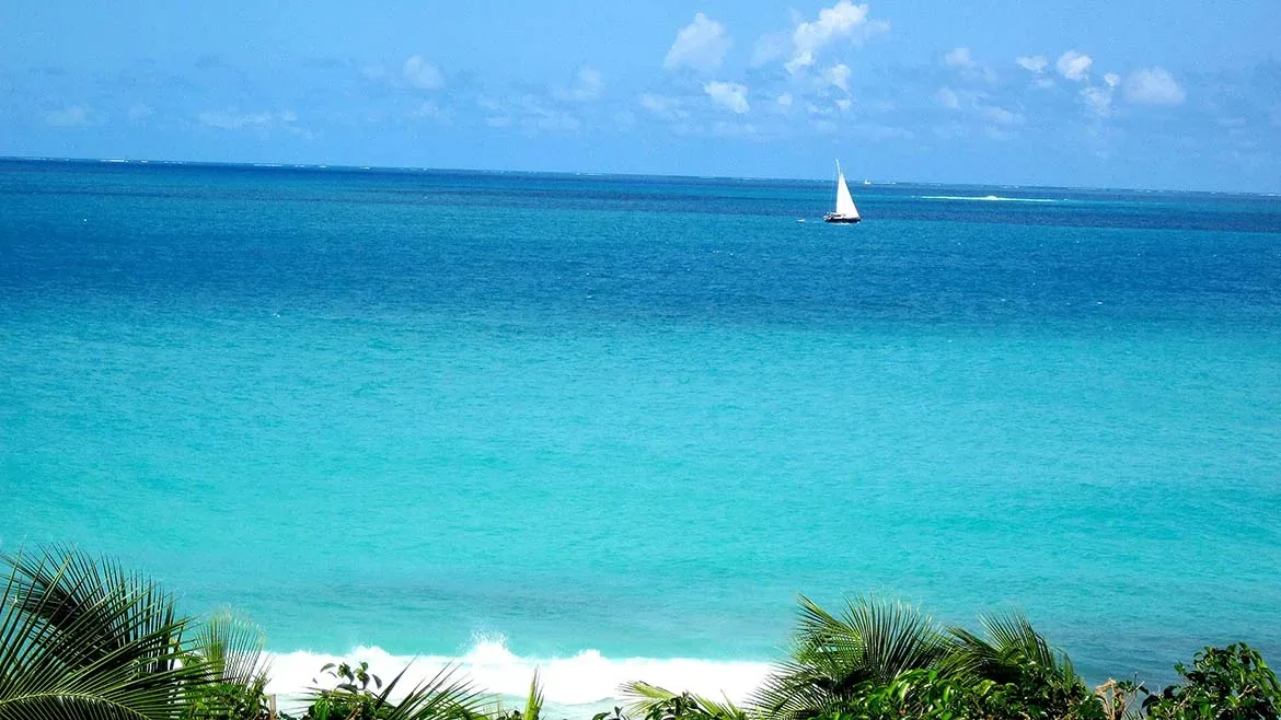 A wide turquoise ocean view with a single white sailboat on the horizon under a blue sky.