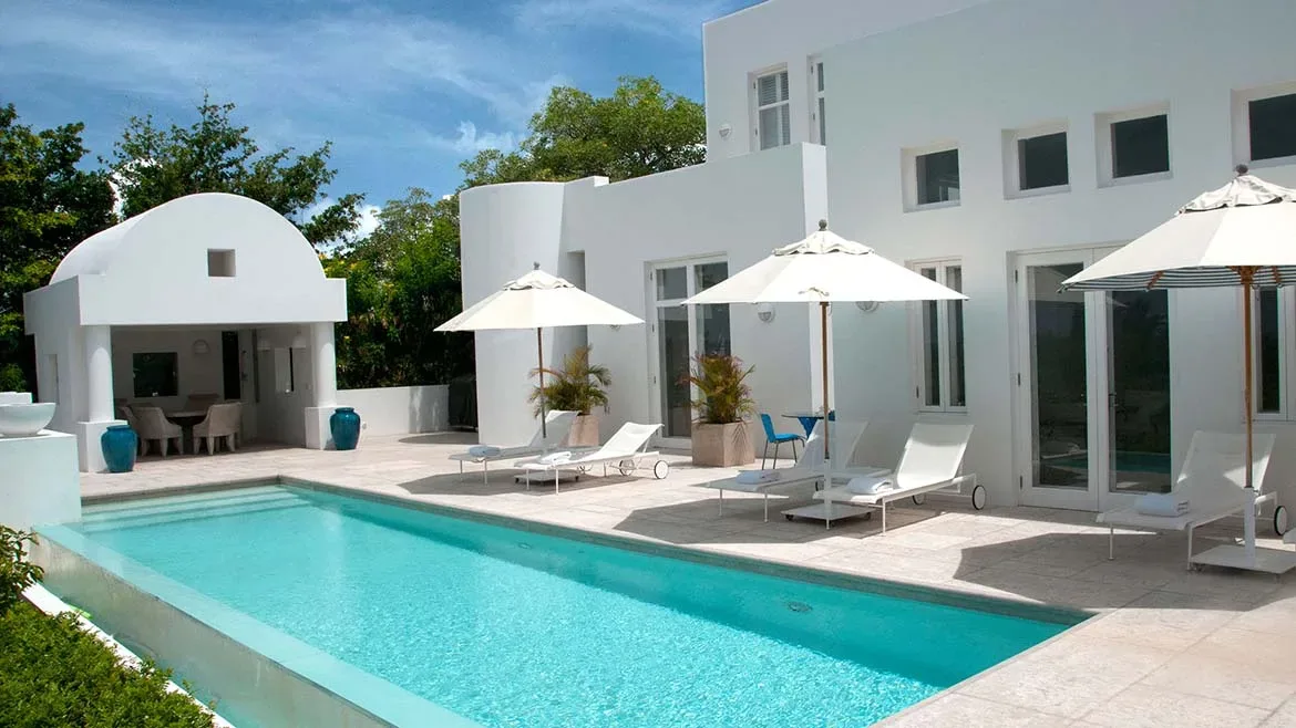 Bright white villa architecture with a rectangular swimming pool, lounge chairs, and white parasols.