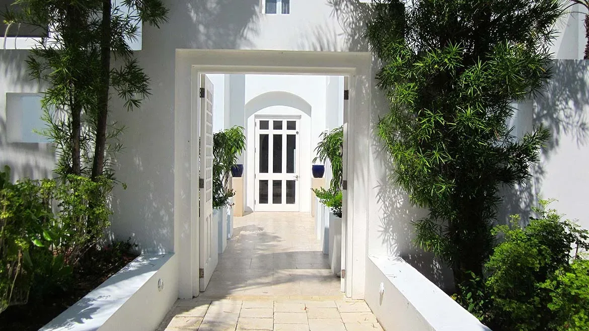 A bright white outdoor corridor with open doors and lush green tropical plants on either side.