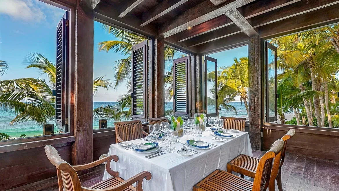Dining table set inside a rustic wooden gazebo with open shutters overlooking palm trees and the ocean.