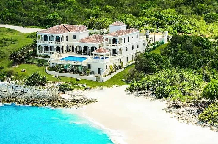 Aerial view of a large white luxury villa with a swimming pool next to a white sand beach and turquoise water.