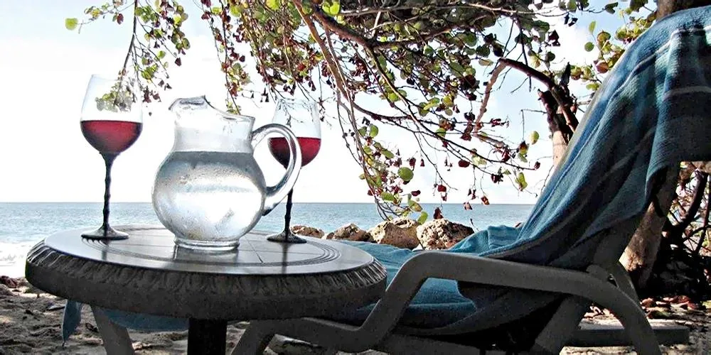 A relaxing beach setup with wine glasses and a water pitcher on a side table next to a lounge chair.