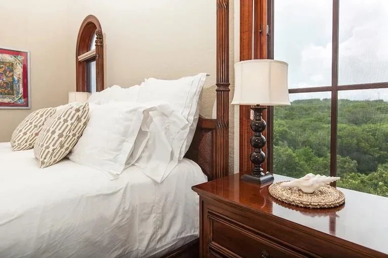 Close-up of a luxury bed with white linens and decorative pillows next to a wooden nightstand with a lamp.