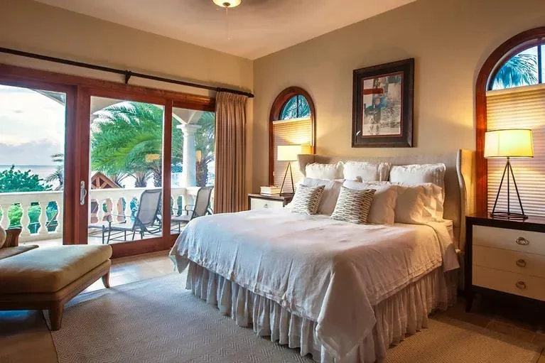 An elegant bedroom suite with soft lighting and a view of the tropical palm trees and ocean through the balcony doors.