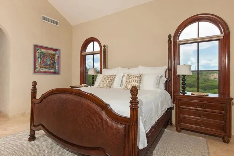 A spacious guest bedroom featuring a large wooden bed, white bedding, and arched windows overlooking the greenery.