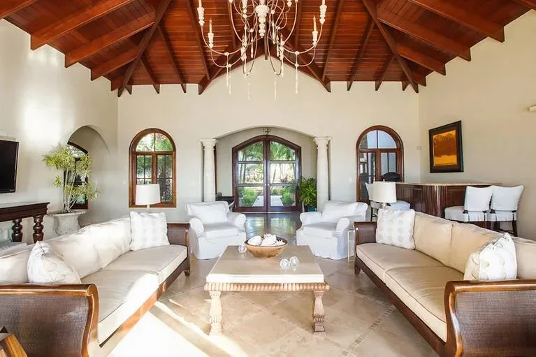 A bright living room arrangement with cream-colored sofas and armchairs centered around a coffee table under a crystal chandelier.