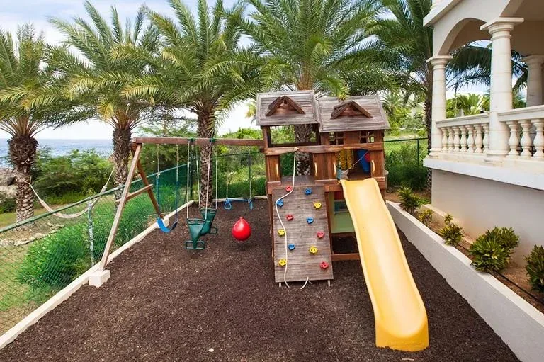 An outdoor children's play area with a wooden playhouse, yellow slide, swings, and a climbing wall set against a backdrop of palm trees.