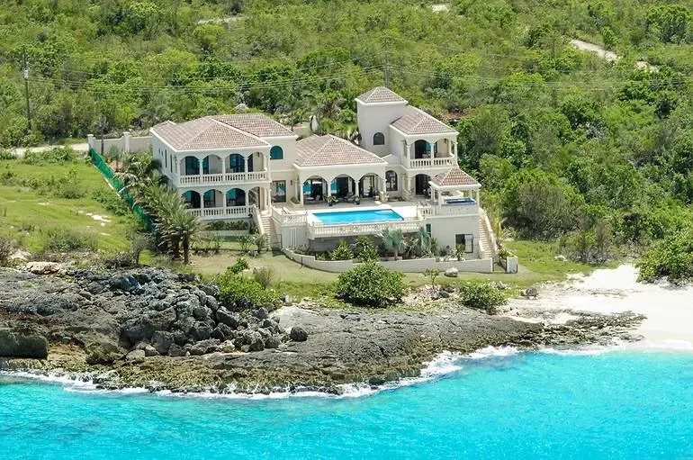 Elevated view of a white villa featuring a pool terrace overlooking a rocky shoreline and clear blue ocean.