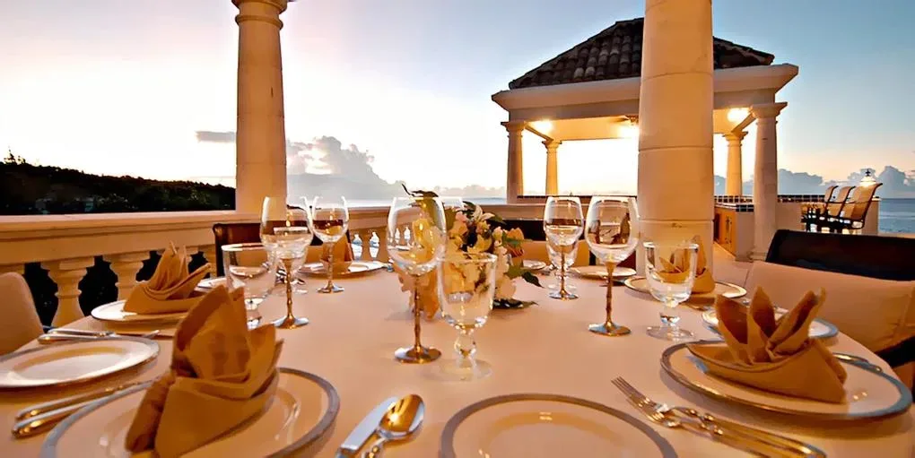 A dinner table set for a sunset meal on the terrace, with the gazebo and ocean visible in the background.