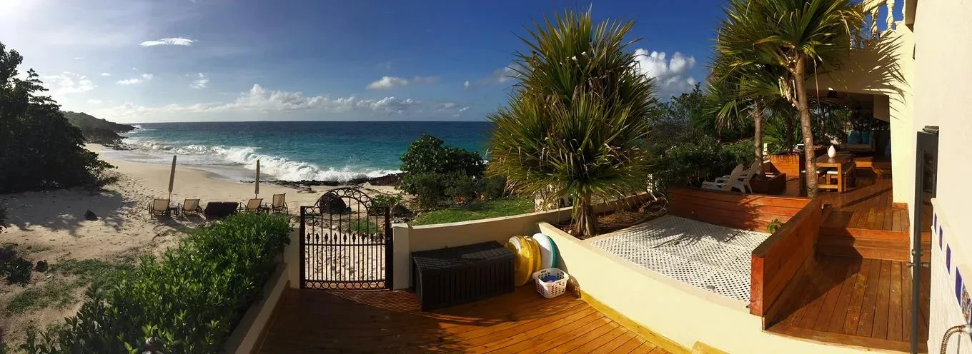 A wooden terrace and tropical garden area featuring palm trees and a black wrought-iron gate leading to the beach.