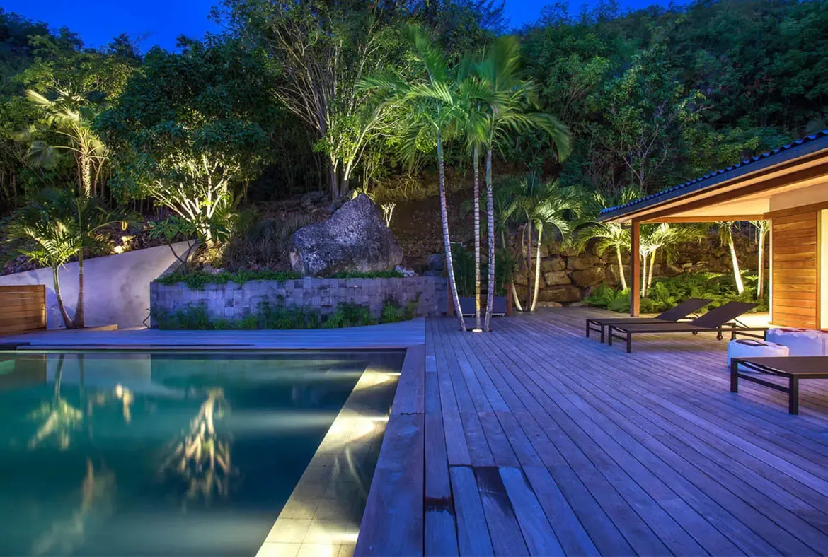 An infinity pool with turquoise water sits before a modern white villa, where white loungers and tropical gardens perch on a lush green hillside beneath a bright sky.