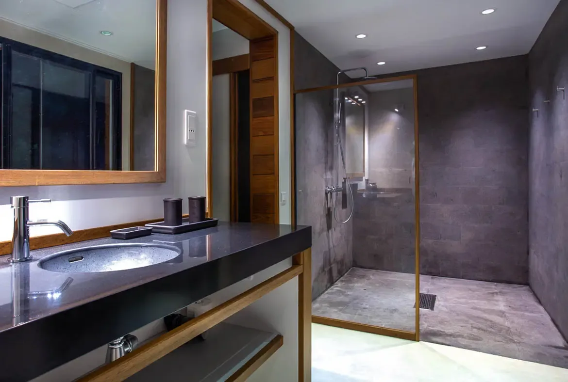 An open-air shower with natural stone walls sits beneath palm trees, where lush tropical plants create a private and tranquil outdoor bathing space.