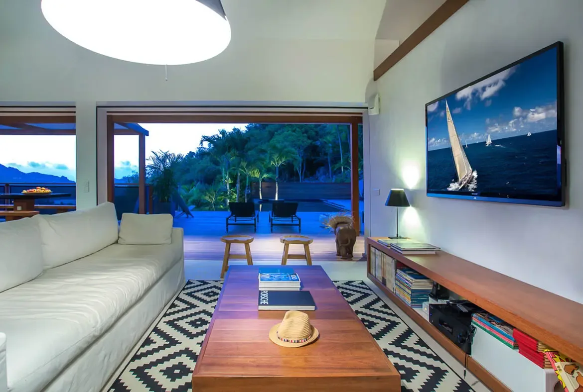 A white bed with plush pillows sits beneath open glass doors, where a private terrace leads to an infinity pool and a turquoise ocean view.
