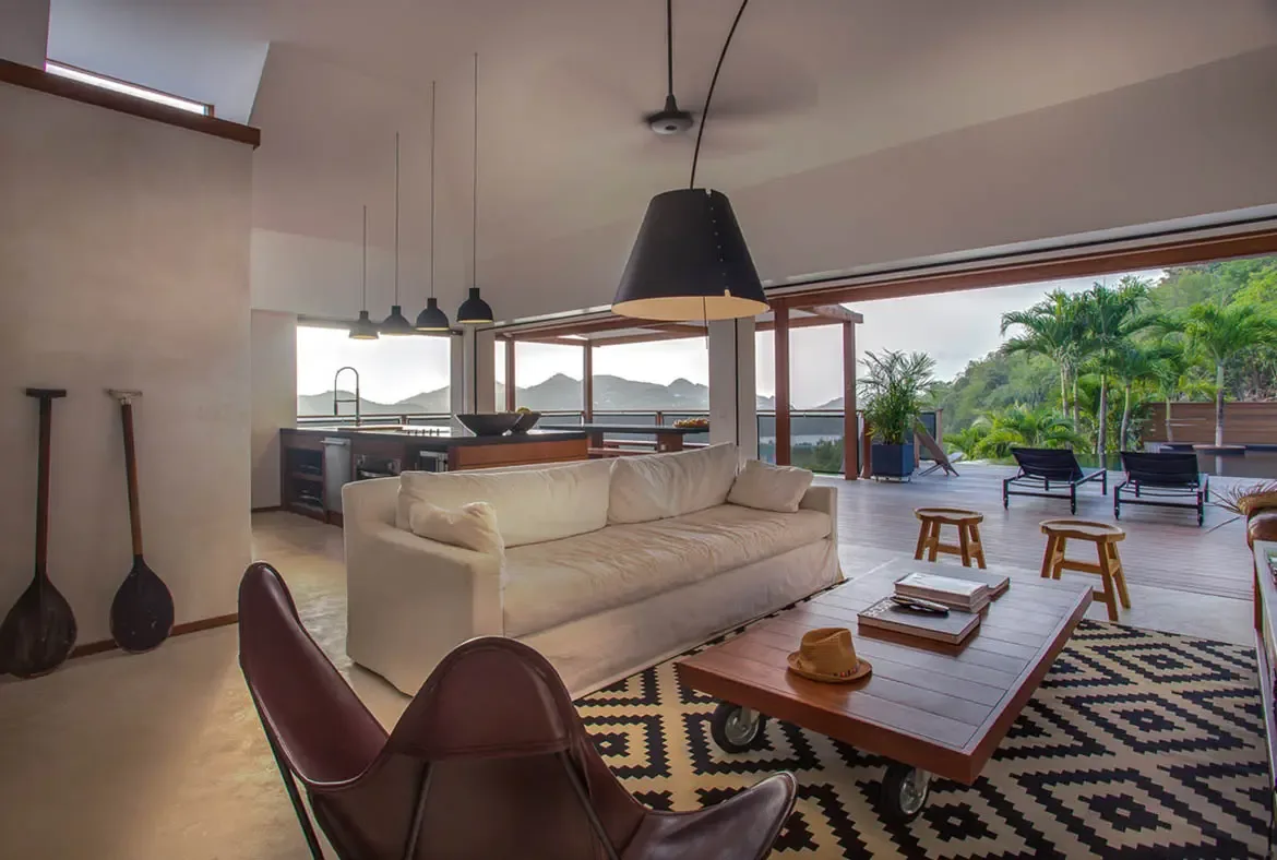 A white kitchen island with bar stools sits before open glass doors, where a sun-drenched terrace and a turquoise ocean fill the space with coastal light.