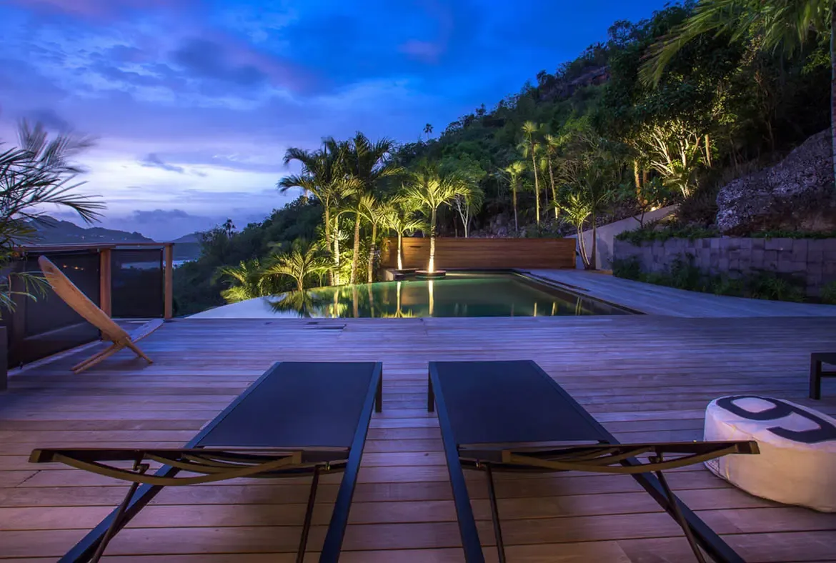 White loungers beside an infinity pool sit on a spacious deck, where a white cabana and palm trees frame a turquoise ocean beneath puffy cumulus clouds.